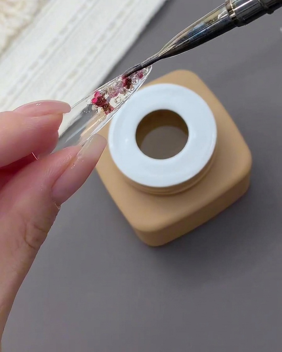 A detail brush applying a thick layer of clear gel over dried flowers and rhinestones encapsulated in the back of a nail tip.