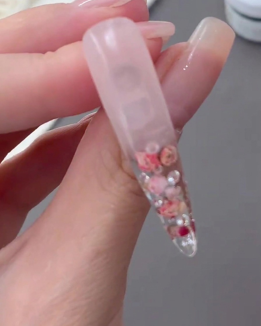 A clear, cured nail tip held up to show the encapsulated floral design and milky gradient background.