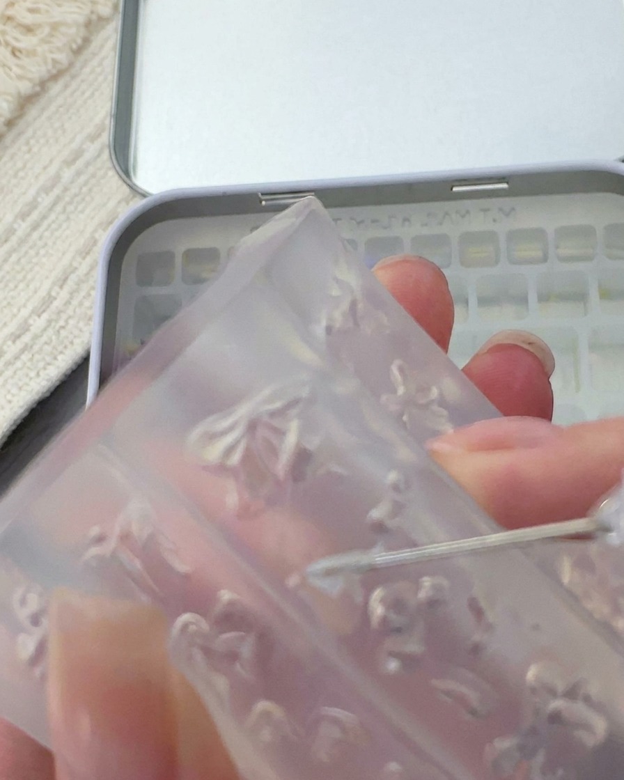 A metal tool lifting a small, delicate white 3D bow from a clear silicone mold.
