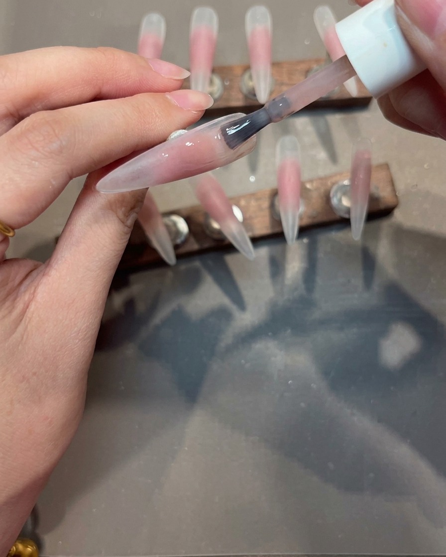 A brush applying a smooth coat of nude gel polish over a nail tip that has a pink blush center.