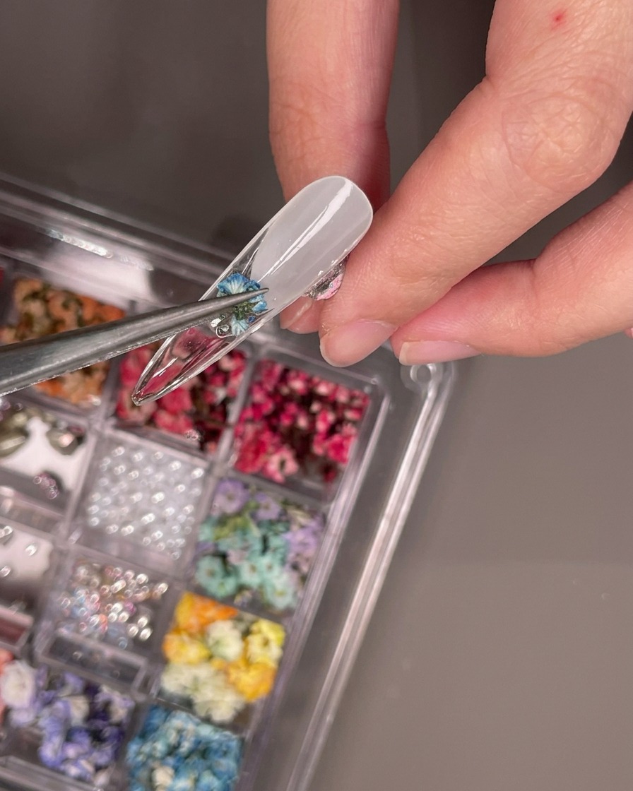 Tweezers placing a small dried blue flower onto the underside of a clear nail tip.