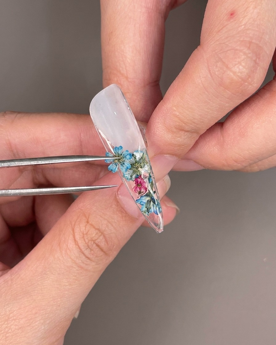 Close-up of clear rhinestones being placed among dried flowers on the underside of a nail tip.