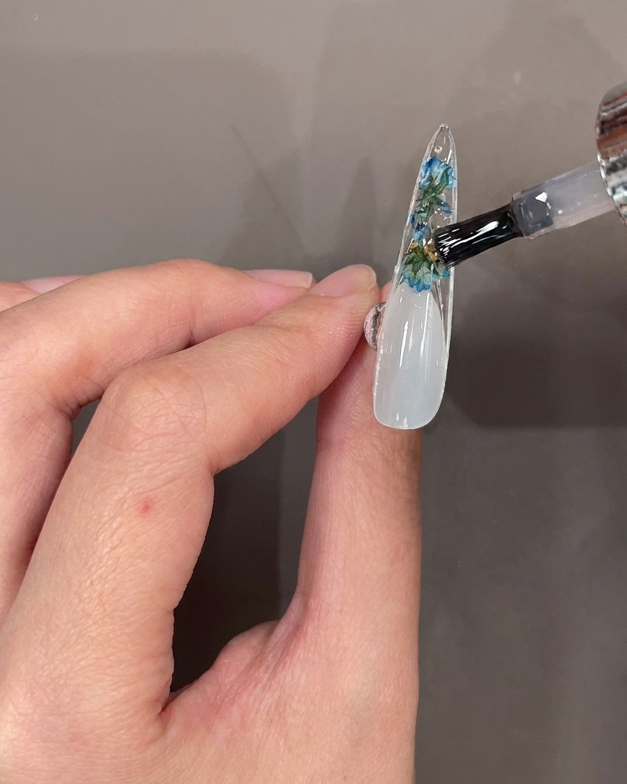 A nail brush applying clear top coat over flowers and stones on the underside of a nail tip.