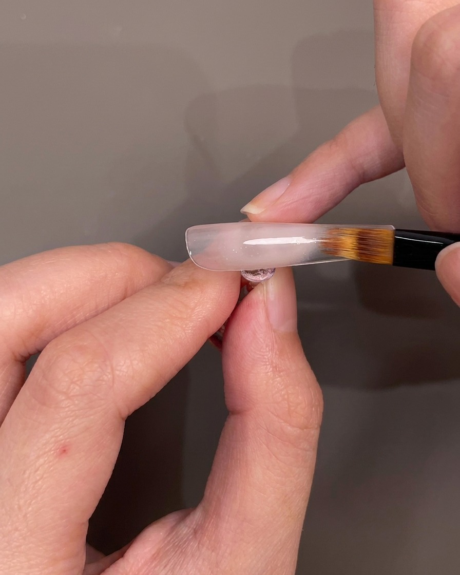 A brush blending nude gel polish downwards on a nail tip to create an ombre effect.