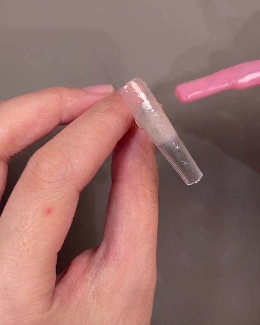 A brush applying sheer pink gel polish diagonally across a nail tip.