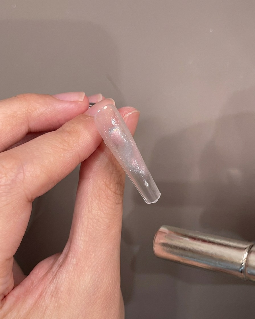 A hand holding a clear nail tip while a cylindrical magnetic wand hovers over the wet cat-eye gel to create a shimmer effect.