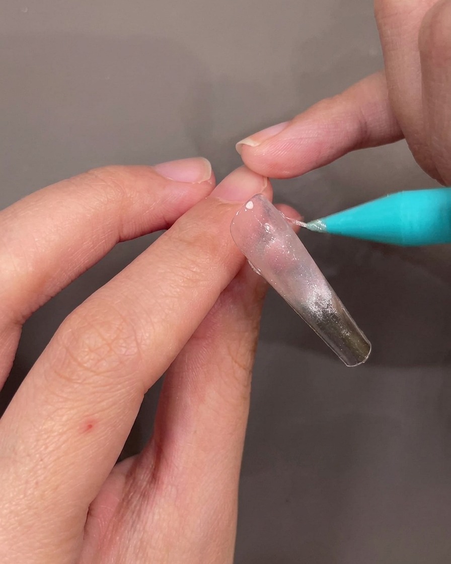 A close-up view of a hand applying a thin, diagonal line of silver glitter gel across a clear nail tip.