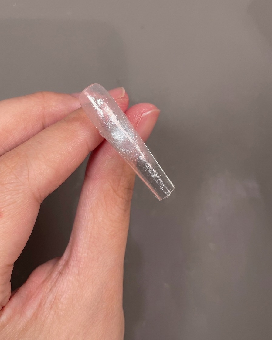The underside of a clear nail tip with a small blue dried flower being carefully placed into a layer of wet gel.