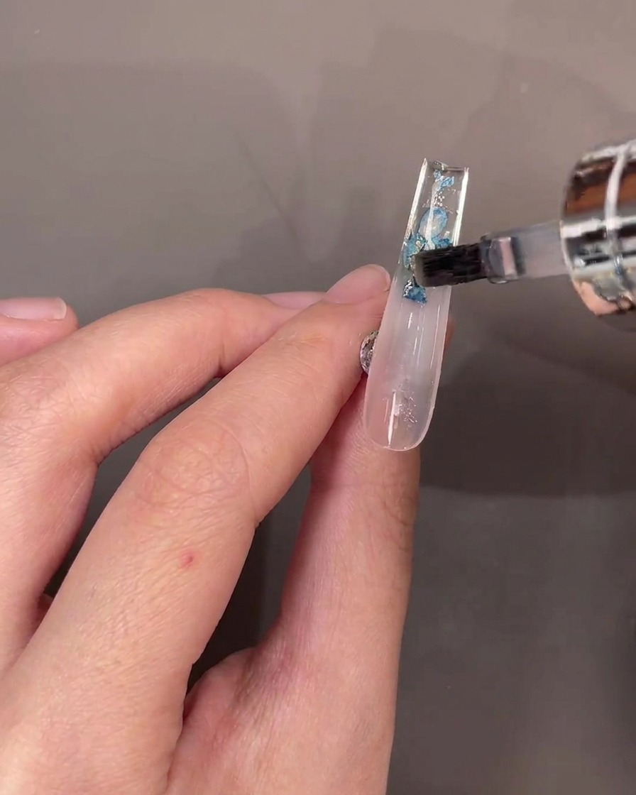 A brush applying a layer of clear top coat over the dried flowers on the underside of a clear nail.