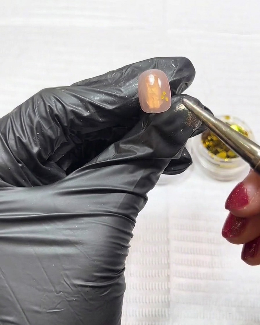 A close-up of a nail showing gold and silver glitter being placed with a tool on a nude gel base.