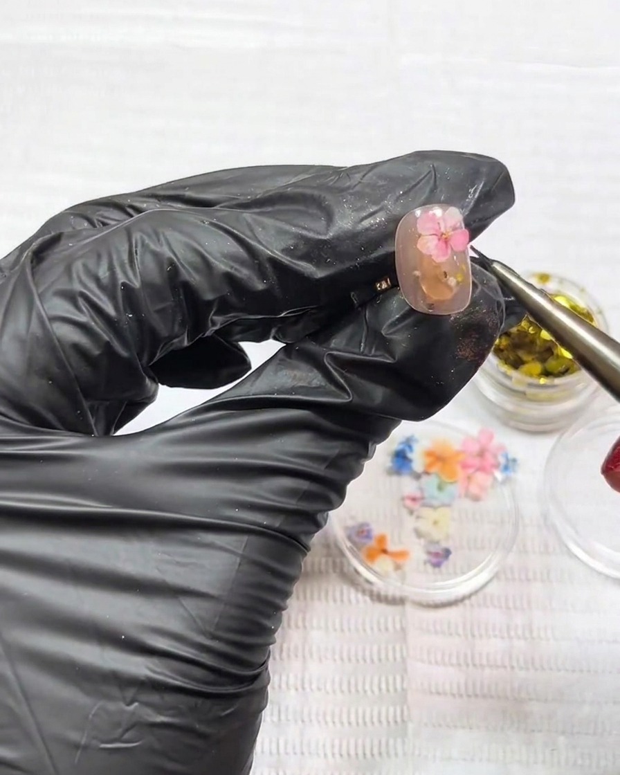 Tweezers placing a small, flat pink dried flower onto the surface of a nail.