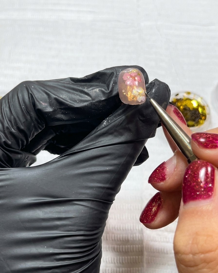 A detail brush applying small, irregular flakes of gold leaf to a nail already decorated with dried flowers.