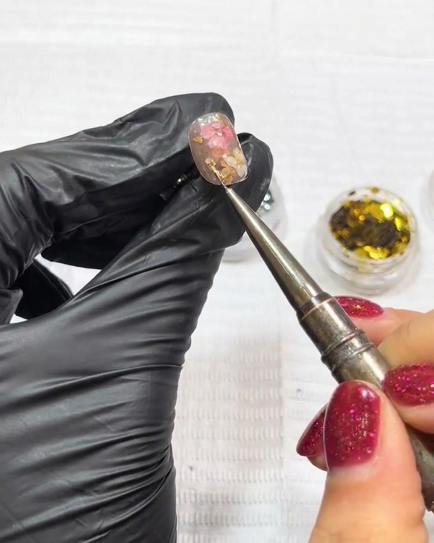 Close-up of tweezers placing a tiny metallic star charm onto a nail decorated with dried flowers and gold leaf.