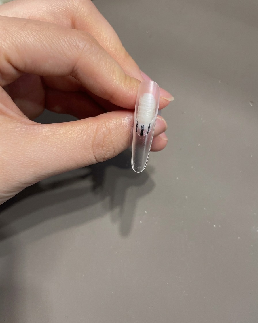 Tweezers placing a small 3D gel bow onto the surface of a manicured fingernail.