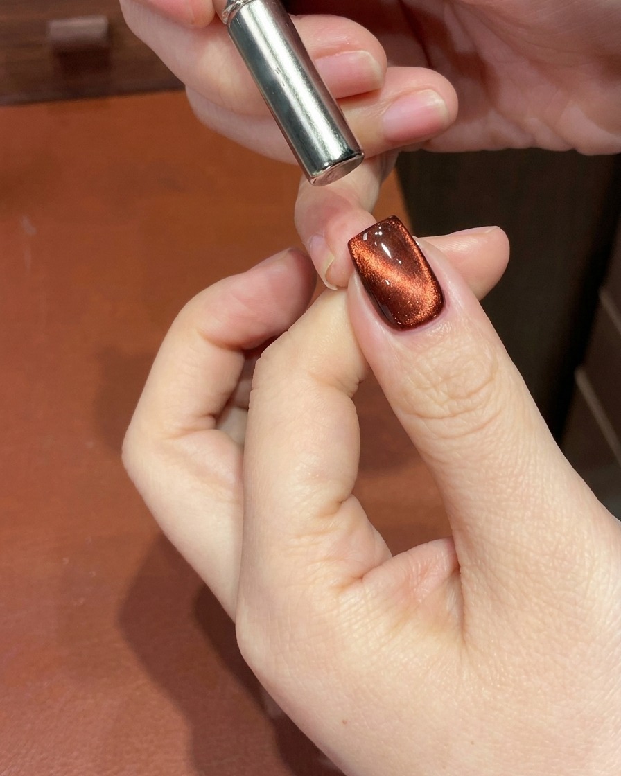 A close-up of a red cat-eye gel nail with a magnet being used near the cuticle to shift the magnetic shimmer into a deep gradient.