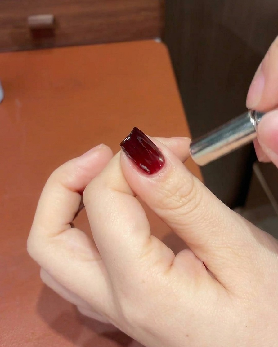 A close-up view of a hand holding a magnetic tool near the tip of a red cat-eye gel nail to refine the shimmer placement.
