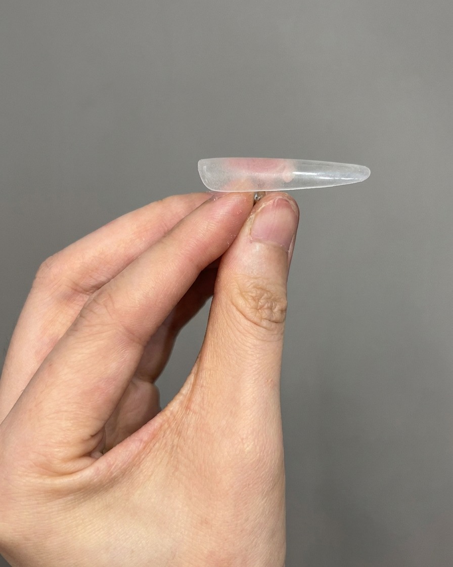 A close-up view of a tool being used to remove fine dust from the surface of a clear nail tip.