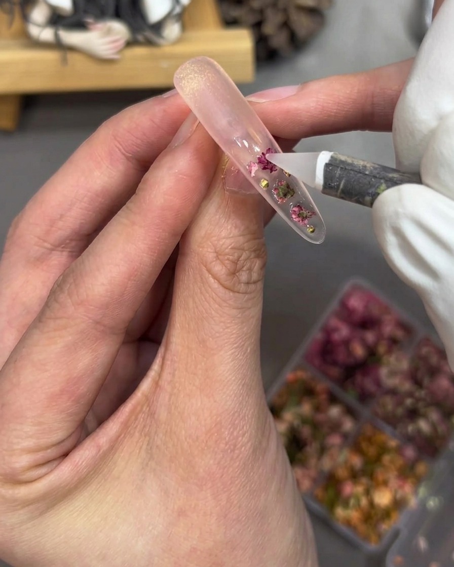 A close-up of a hand using a tool to place small gold pearls and pink rhinestones onto a clear gel-coated nail tip.