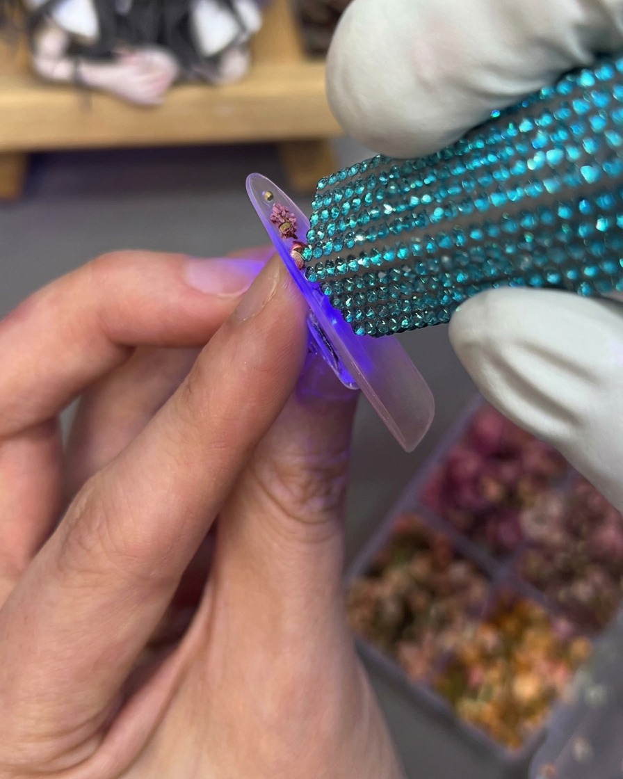 A hand holding a mini UV lamp over a nail tip that has dried flowers and rhinestones embedded on the surface.