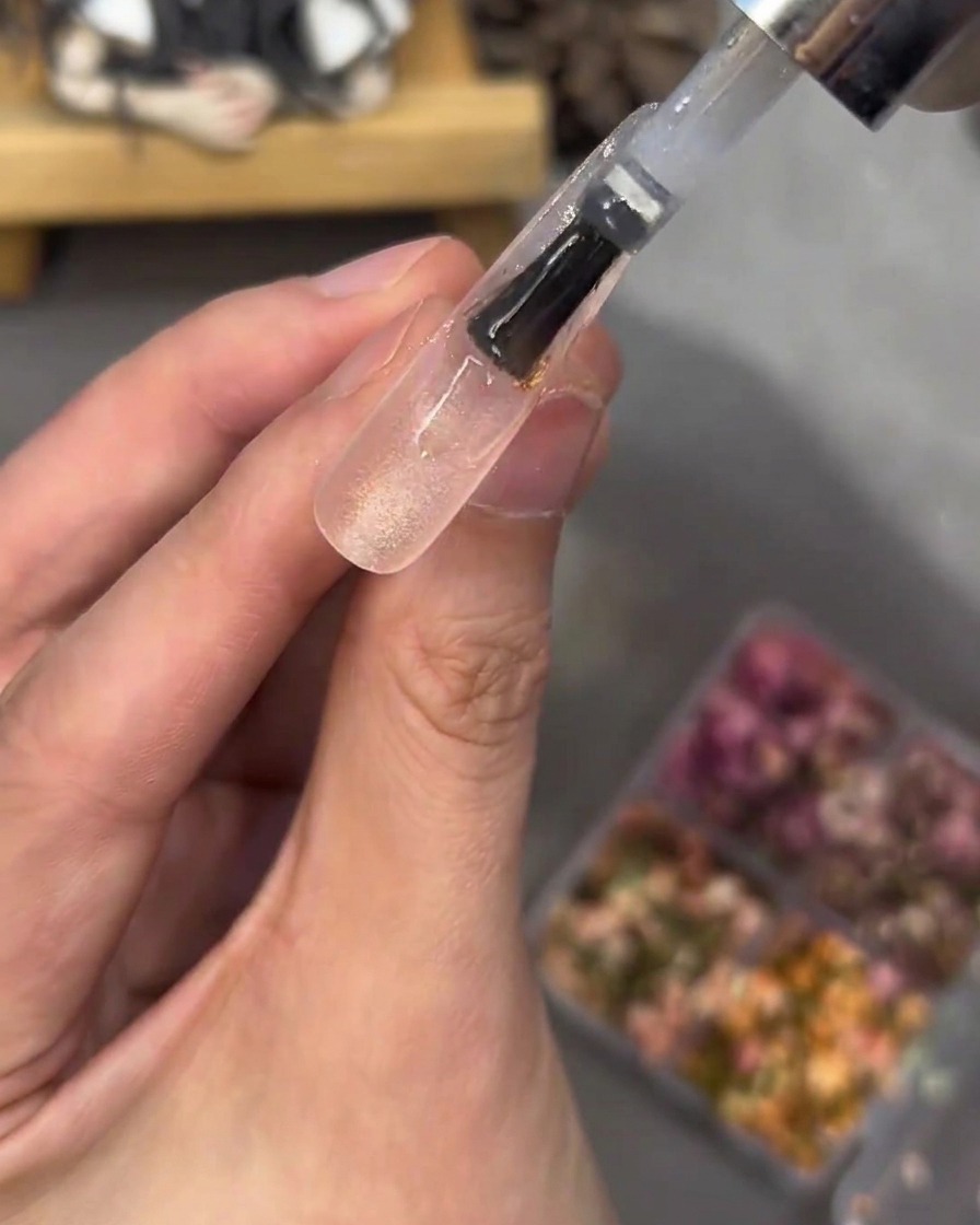 A brush applying a clear, thick layer of gel over a nail tip embedded with flowers and rhinestones, smoothing the surface.