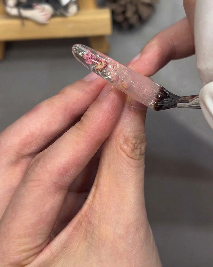 A brush applying clear top coat gel to a fully decorated nail tip, creating a glossy finish.