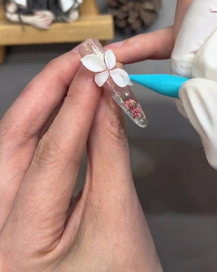 A detail shot showing a brush applying a soft pink tint to the white sculpted petals on the nail.