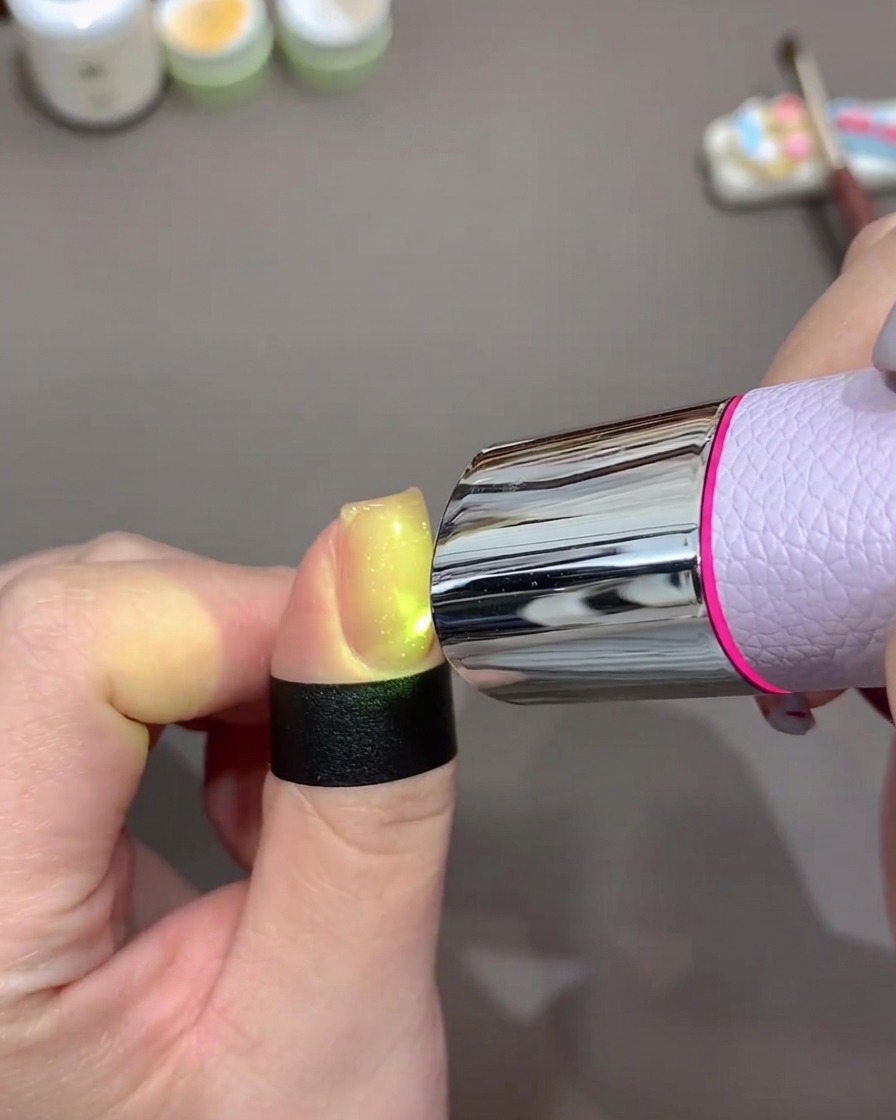 A fingernail glowing brightly under a small, portable purple UV/LED lamp to cure the gel polish.