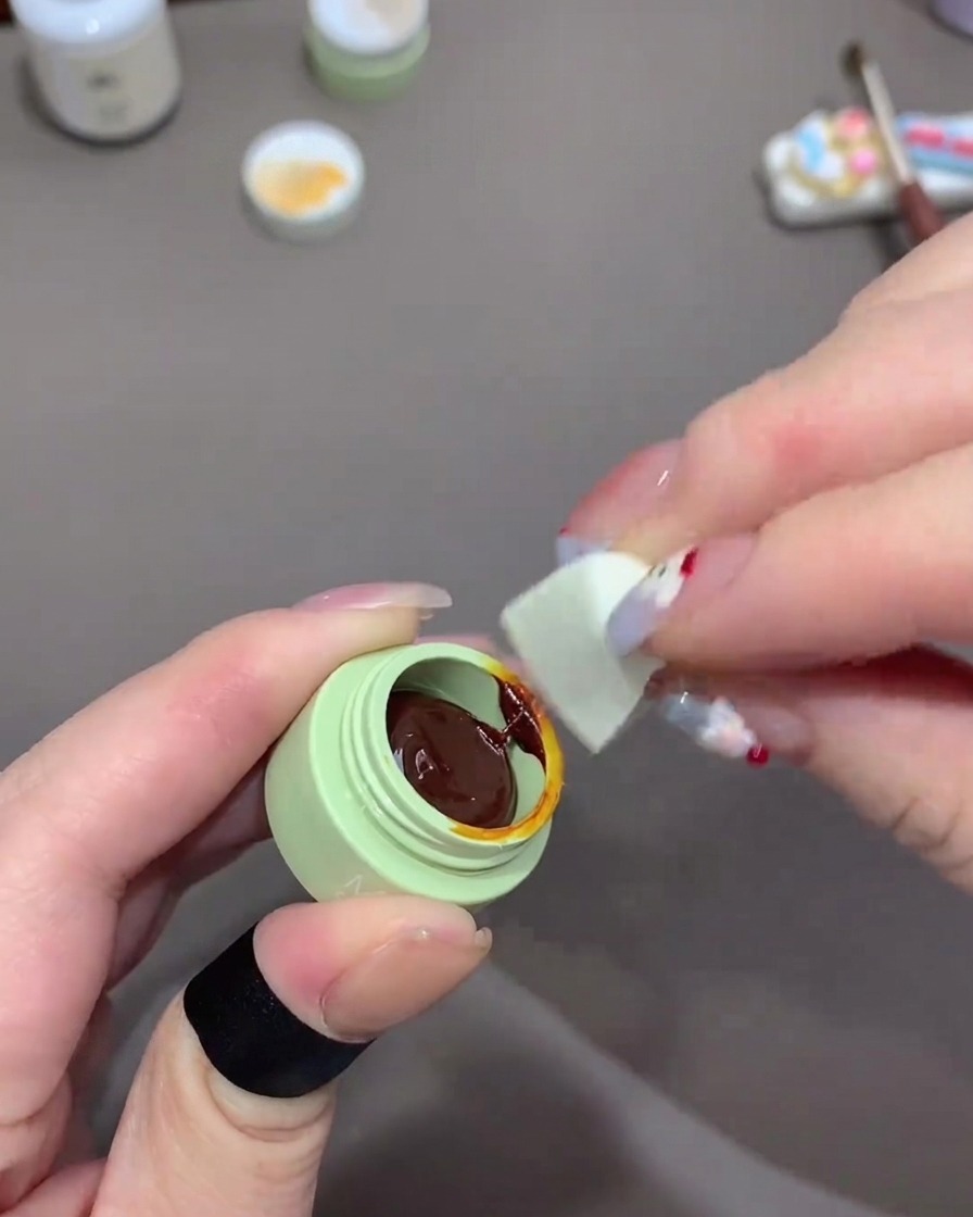 A hand using a small piece of a white makeup sponge to pick up dark orange gel polish from a small green pot.