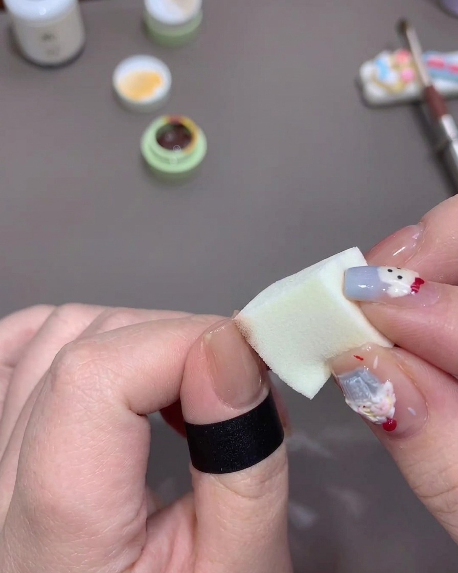 A hand dabbing a small makeup sponge loaded with orange gel onto the tip of a thumb nail to create a soft gradient fade.