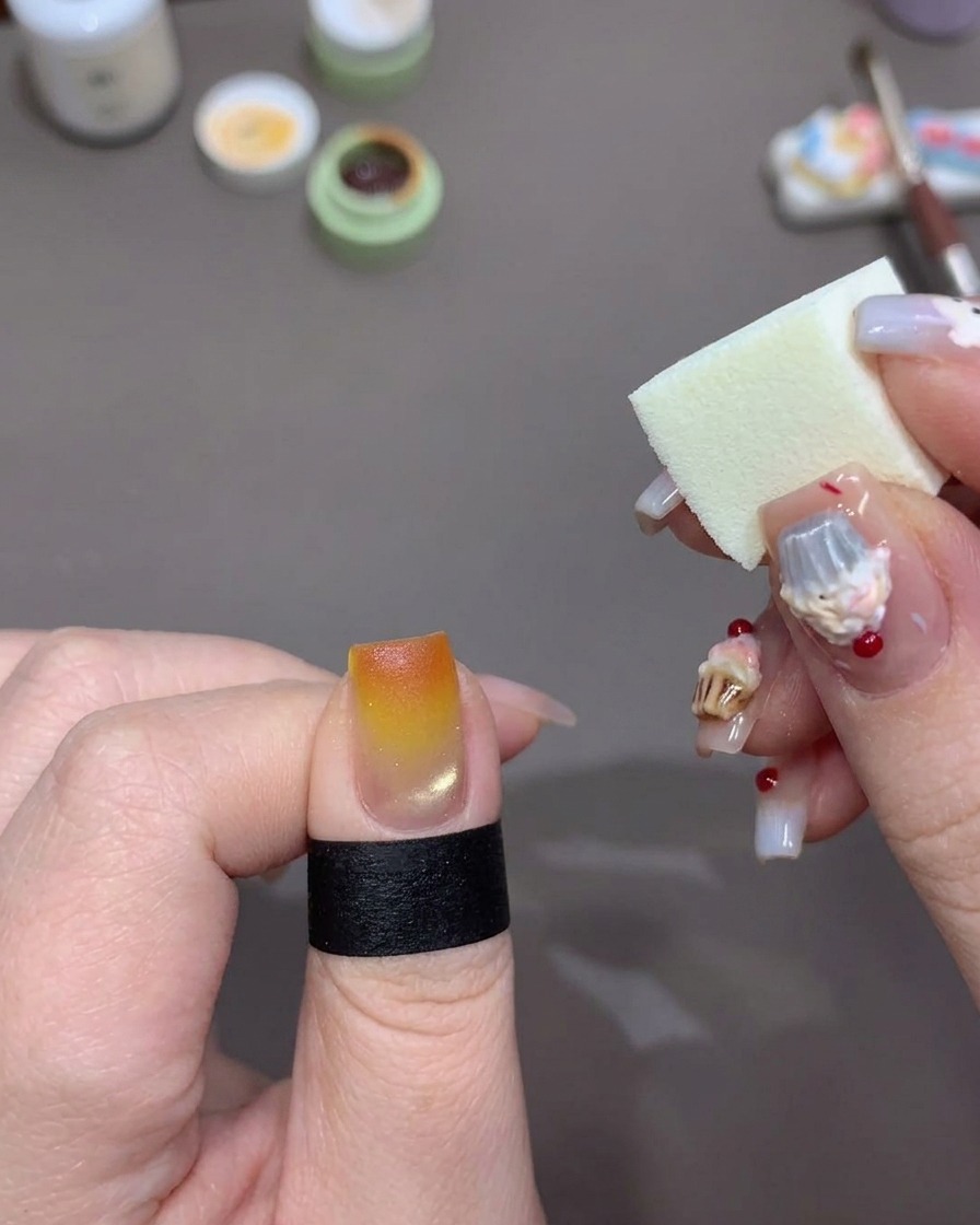 A white makeup sponge dabbing bright orange gel polish onto the very tip of a fingernail to intensify the sunset gradient effect.