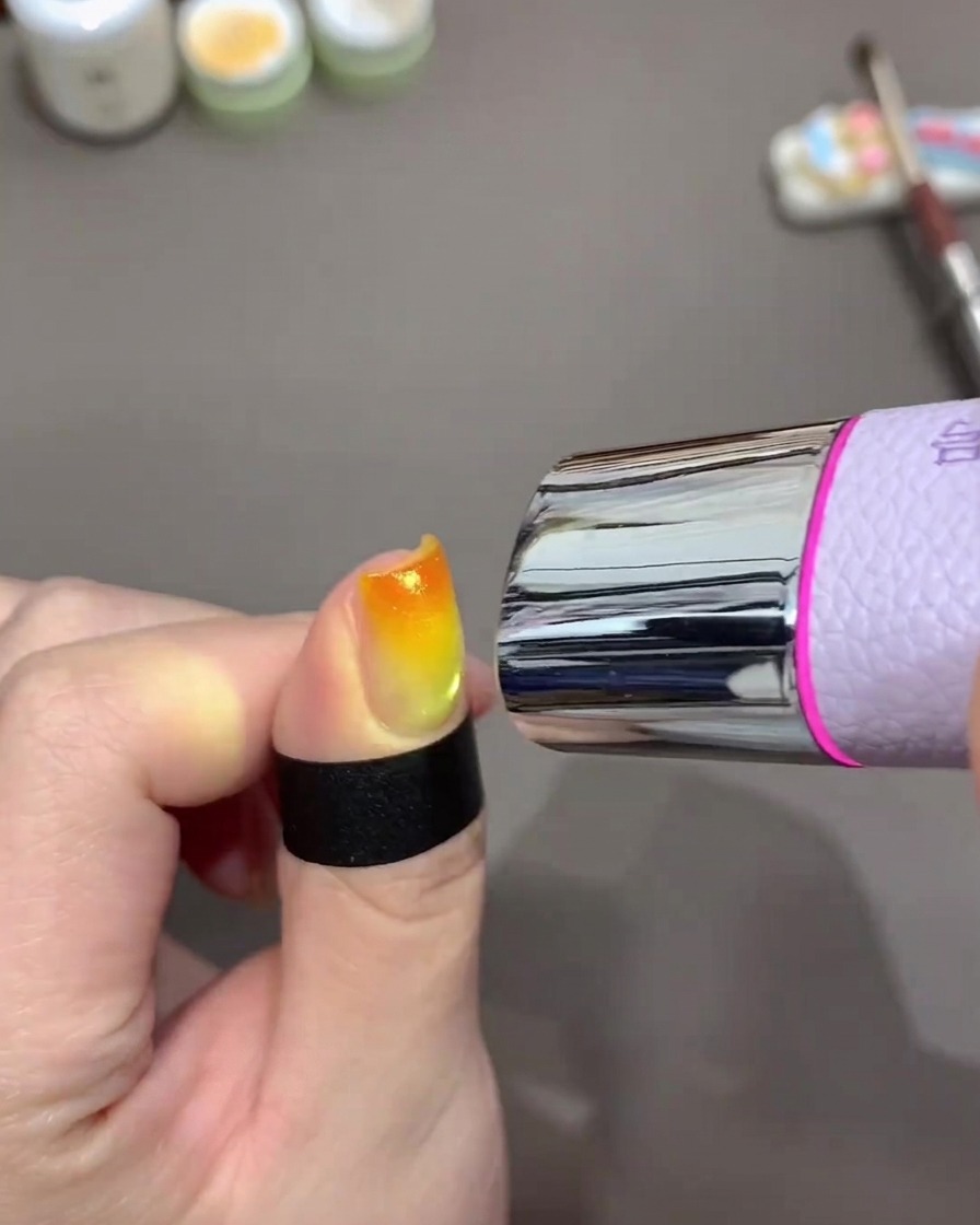 A mini handheld UV lamp positioned over a fingernail to cure a bright orange and yellow gradient gel design.