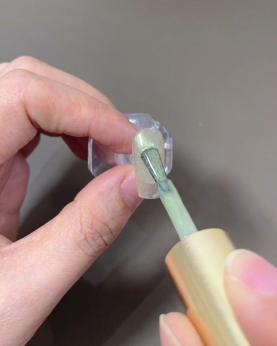 A nail brush applying light-colored glass cat-eye gel to a nail.