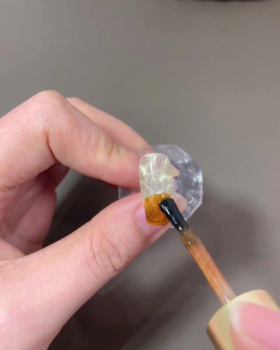A brush applying amber-colored translucent gel onto the tip of a nail.