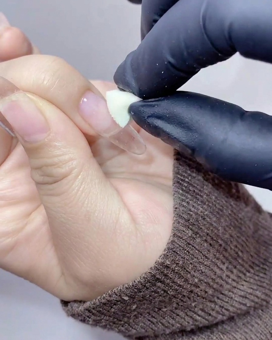 Gloved hands using a small cosmetic sponge wedge to dab peachy-orange gel onto the center of a clear nail extension.