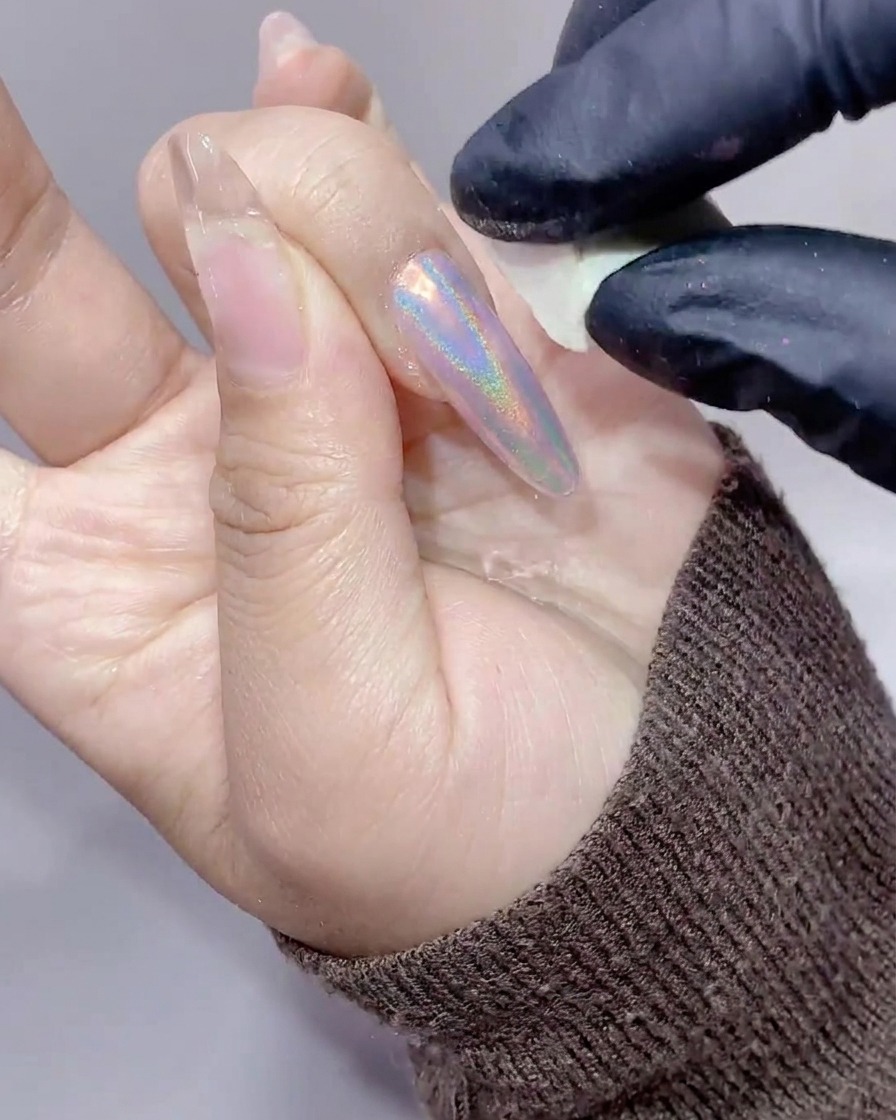 A close-up of a hand with long nails being wiped clean of excess aurora powder using a small white makeup sponge.
