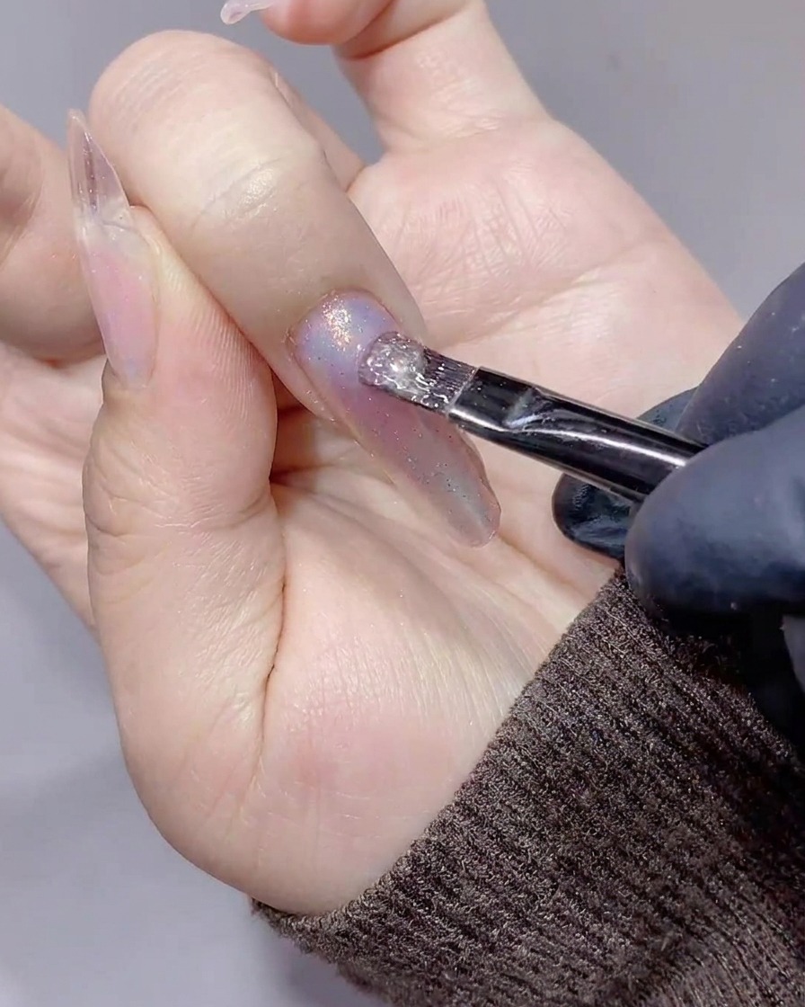 A brush applying a clear, shimmering magnetic cat-eye gel onto a nail already decorated with aurora powder.