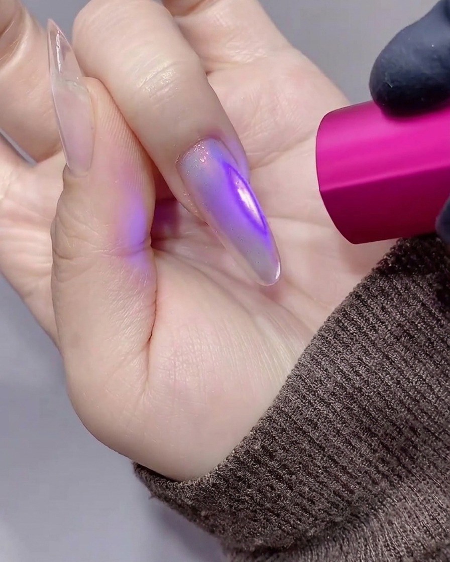 A close-up of a hand with a long, translucent purple-toned stiletto nail being cured under a small handheld pink UV/LED lamp.