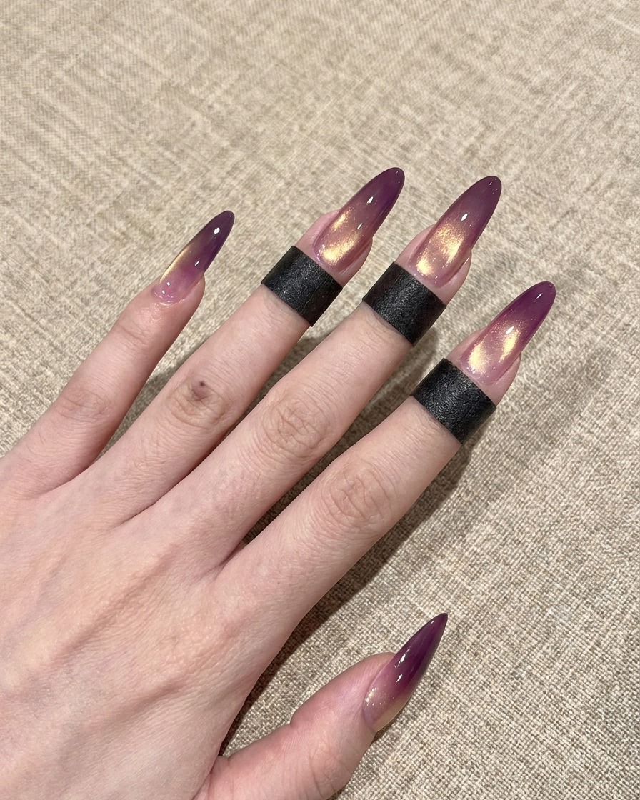 A close-up of a brush applying a glossy clear top coat over a purple and gold gradient cat-eye nail.