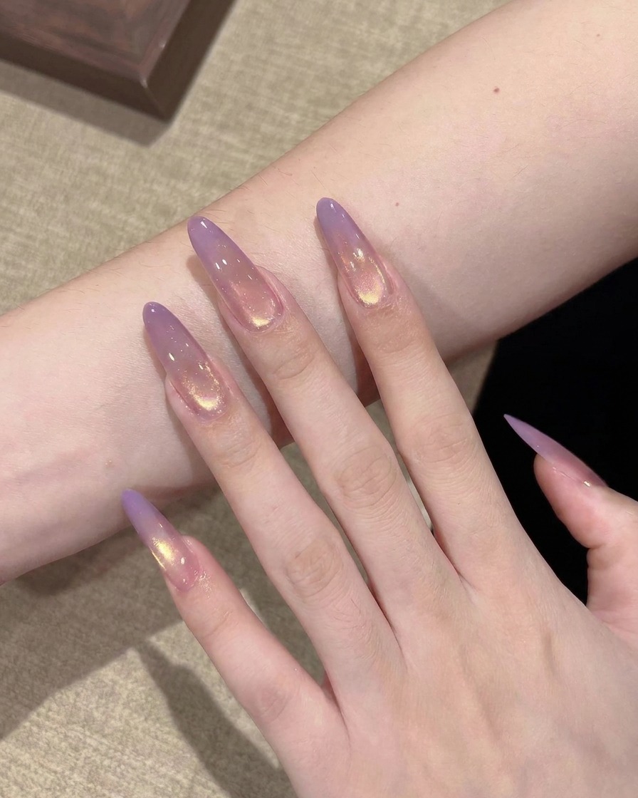A close-up view of a flat brush depositing a small bead of translucent purple gel onto the tip of a clear, stiletto-shaped nail extension.