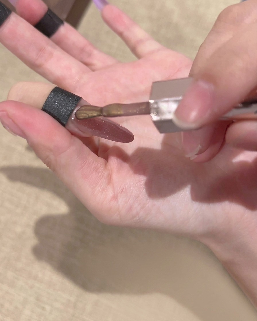 A brush from a gel polish bottle applying a layer of gold magnetic cat-eye gel over a nail with an existing purple gradient.