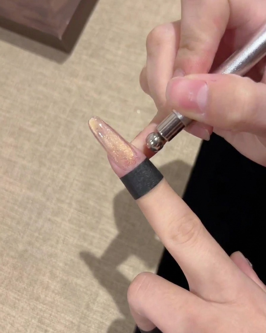 A close-up view of a metal magnet tool being held horizontally over a nail to manipulate magnetic gel particles.