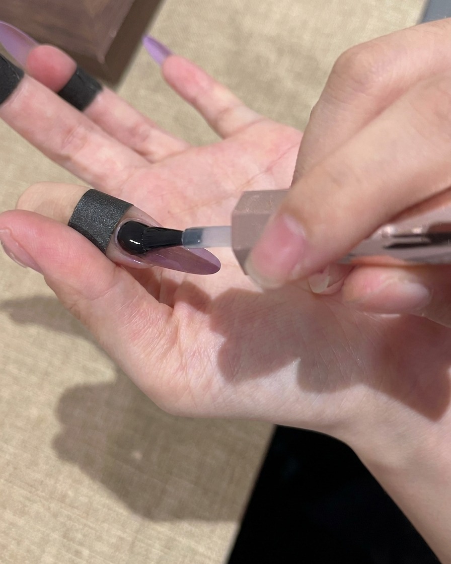 A brush applying a clear, glossy top coat over a purple and gold cat-eye nail.