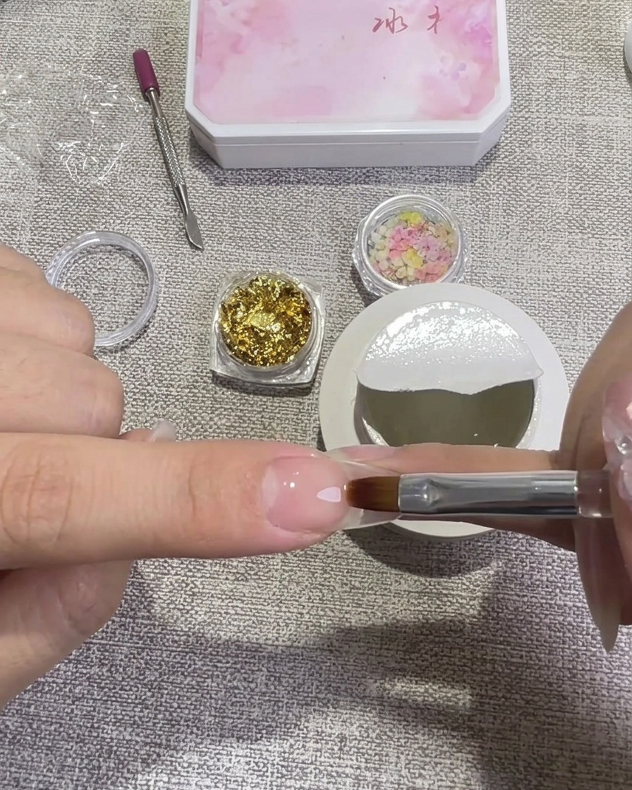A hand using a flat gel brush to apply a layer of clear hard gel over a prepped nail, with small jars of gold foil and dried flowers in the background.