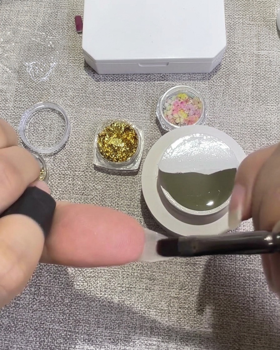 Tweezers placing small, colorful dried flower petals onto a nail covered in wet, clear gel.