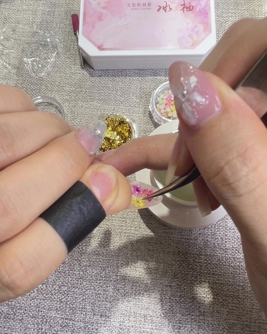 Tweezers delicately repositioning a dried flower petal on a nail surface to achieve a balanced design.