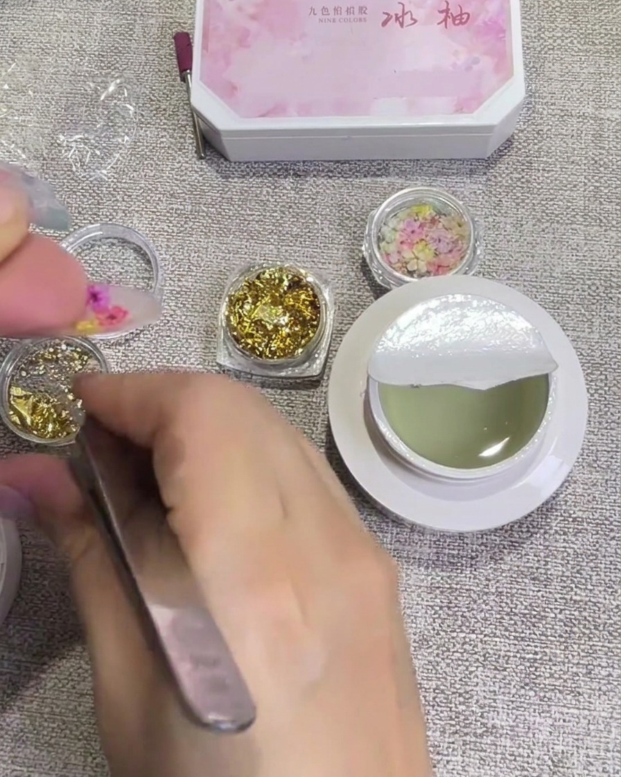 Small gold foil flakes being applied to the nail surface near the dry flowers using tweezers.