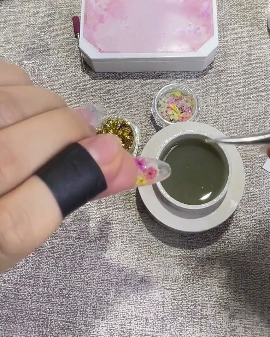 A close-up view of tweezers placing a small, shimmering piece of gold leaf onto a clear, gel-coated nail surface.