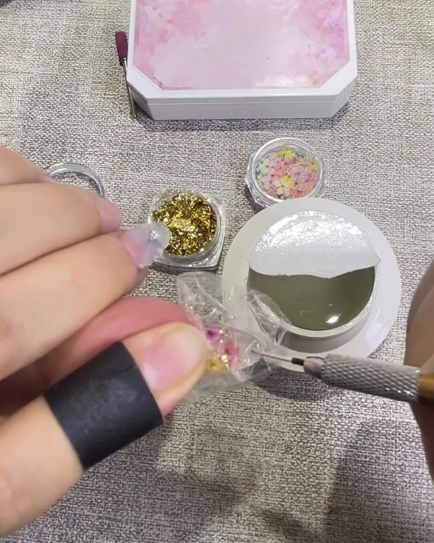 A brush applying a glossy, clear top coat over a nail decorated with pressed flowers and gold foil, making it look wet and translucent.