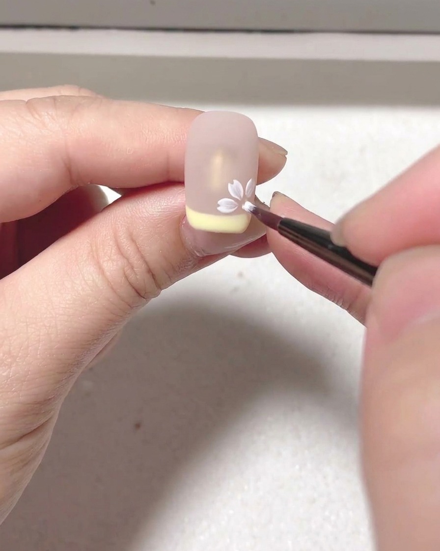 A close-up view of a brush applying white blooming gel to create a petal shape on a nude nail surface.