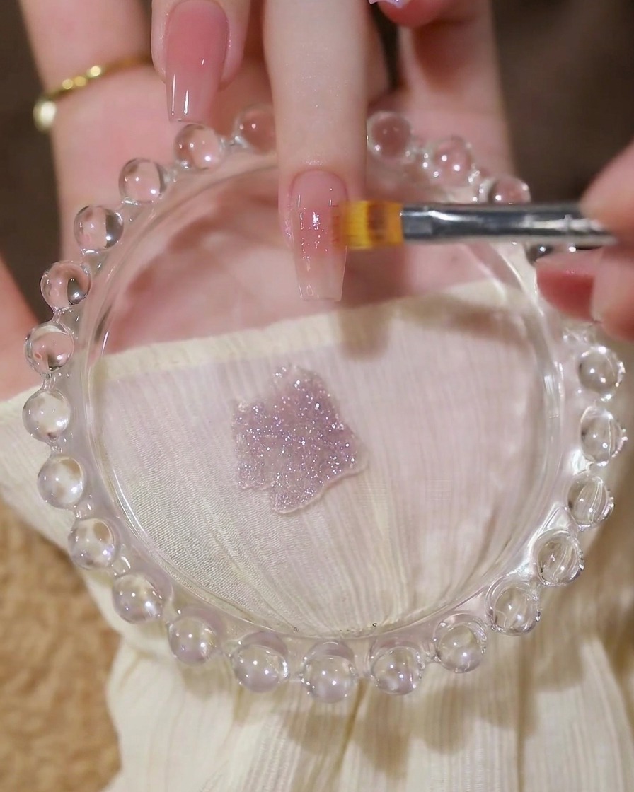 A brush applying a layer of sheer, glittery sequin gel over a nude nail base.